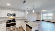 This single-floor residence offers a well-designed, contemporary layout with a clear separation between living, sleeping, and utility spaces. The kitchen serves as a focal point, showcasing white shaker-style cabinetry extending to the ceiling, granite countertops, and a full suite of stainless steel appliances including a refrigerator, range, microwave, and dishwasher. A peninsula with an undermount sink and pendant lighting overhead creates a natural divide between the kitchen and the open living area, which features wood-look plank flooring that runs throughout the main spaces.

The living area is generously proportioned and flows seamlessly from the kitchen, with neutral walls and access to what appears to be a balcony through sliding glass doors covered by vertical blinds, allowing for natural light and outdoor views. The bedroom is carpeted for comfort and includes a window for natural light. An adjacent full bathroom features a single-sink vanity with a large mirror, a toilet, and a tub/shower combination, all finished in a clean white palette with wood-look flooring. A spacious walk-in closet with wire shelving provides ample storage. Dark baseboards throughout offer a modern contrast to the light walls and flooring.