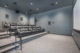 Located at 2015 E Springs Dr in Madison, Wisconsin, this purpose-built private screening room delivers a true cinematic experience in a compact, thoughtfully designed space. The room is finished throughout in deep charcoal tones — from the dark-painted walls and ceiling to the matte trim — creating a fully immersive, distraction-free viewing environment. A large, framed projection screen anchors the front wall, providing a theater-quality display surface that commands attention upon entry. Seating is arranged in two tiered rows of plush, black leather reclining chairs, each equipped with built-in cup holders. The elevated rear row ensures unobstructed sightlines from every seat, replicating the stadium-style layout of a commercial cinema. A surround sound speaker system is strategically mounted across the walls at multiple heights, delivering rich, enveloping audio throughout. Recessed ceiling spotlights provide controlled ambient illumination, while subtle wall sconces add a refined accent. Soft, neutral carpet covers both seating tiers, contributing to the room's acoustic dampening. Metal handrails guide movement between levels, and a clearly marked exit door ensures safe, convenient access. This screening room offers a complete, professional-grade home theater experience.