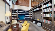 Located at 317 North Broad Street in Philadelphia, this distinguished library lounge is a single-floor living space that masterfully blends classic architectural grandeur with thoughtful, contemporary furnishings. The room commands attention with its dramatically high ceilings, richly paneled woodwork, and ornate decorative columns that speak to the building's historic character.

Floor-to-ceiling built-in bookshelves line the walls, filled with an extensive collection of books and tasteful decorative objects, creating an atmosphere of intellectual warmth. A standout feature is the tufted teal velvet reading nook recessed within the shelving — an inviting alcove accented with plush cushions, perfect for focused reading. An integrated built-in desk with adjustable sconce lighting and paired ottomans offers a dedicated workspace within the shelving unit.

The seating arrangement includes vibrant yellow armchairs, a neutral patterned sofa, and cream accent chairs, all centered around wooden coffee tables to encourage conversation and relaxation. Tall windows fitted with roller shades allow natural light to filter into the space while maintaining a serene ambiance. A direct entrance with glass-paned French doors connects the lounge to an outdoor area, enhancing accessibility and airflow. The carpeted floors and warm lighting complete the refined, welcoming character of this exceptional space.