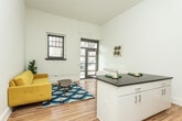 This single-floor unit showcases a compelling mix of industrial heritage and contemporary design. The living area greets visitors through a glass-paneled entry door that floods the space with natural light, complemented by a multi-pane window set against a white-painted exposed brick accent wall — a standout architectural detail that lends the space notable character and texture. Warm hardwood floors run throughout, unifying each area cohesively.

The open kitchen is efficiently laid out with dark countertops, white cabinetry, stainless steel appliances including a refrigerator and dishwasher, and pendant lighting overhead. A generous island with matching dark countertop provides additional prep and dining space.

The bedroom is spacious and airy, benefiting from tall ceilings and clean white walls. Ample built-in cabinetry offers generous storage, and a stacked washer and dryer unit is conveniently located within the room. The bathroom features classic white subway tile in the shower alongside a distinctive patterned mosaic tile floor, adding visual interest to the clean, minimal design.

Overall, the space balances raw architectural elements with polished modern finishes, creating a functional and characterful environment.