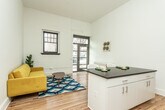 This single-floor unit showcases a compelling mix of industrial heritage and contemporary design. The living area greets visitors through a glass-paneled entry door that floods the space with natural light, complemented by a multi-pane window set against a white-painted exposed brick accent wall — a standout architectural detail that lends the space notable character and texture. Warm hardwood floors run throughout, unifying each area cohesively.

The open kitchen is efficiently laid out with dark countertops, white cabinetry, stainless steel appliances including a refrigerator and dishwasher, and pendant lighting overhead. A generous island with matching dark countertop provides additional prep and dining space.

The bedroom is spacious and airy, benefiting from tall ceilings and clean white walls. Ample built-in cabinetry offers generous storage, and a stacked washer and dryer unit is conveniently located within the room. The bathroom features classic white subway tile in the shower alongside a distinctive patterned mosaic tile floor, adding visual interest to the clean, minimal design.

Overall, the space balances raw architectural elements with polished modern finishes, creating a functional and characterful environment.