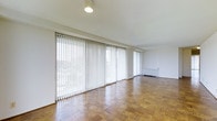 This single-floor space presents a clean, open layout defined by warm parquet wood flooring arranged in a classic block pattern throughout. The most striking feature of the main living area is the expansive wall of floor-to-ceiling windows dressed with vertical blinds, allowing for adjustable natural light and views of the surrounding greenery. The generous window coverage creates a bright, airy atmosphere while offering a strong connection to the outdoors. The interior walls are finished in a crisp white, contributing to the space's understated, neutral palette. Ceiling-mounted lighting provides functional illumination throughout. A baseboard heating unit runs along the exterior wall, suggesting efficient, zone-controlled climate management. The open floor plan allows for flexible furniture arrangement, making the space adaptable to a variety of uses. Overall, the space delivers a straightforward, functional design with classic material choices and an abundance of natural light as its defining characteristic.