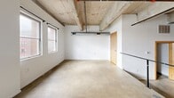 Located at 281 5th Street East in downtown Saint Paul, Minnesota, this fourth-floor loft-style unit showcases an industrial aesthetic rooted in its original building structure. Polished concrete floors extend throughout the entire space, complementing the exposed concrete ceiling beams and visible ductwork overhead — hallmarks of a converted urban building. The open main area is bright and functional, with white walls offering a clean contrast to the raw architectural elements. Natural light filters in through multiple windows, contributing to an airy feel despite the compact footprint. The kitchen is efficiently arranged along one wall, featuring warm wood cabinetry, dark countertops, a double sink, an electric range, and a refrigerator. A pendant light fixture adds character to the kitchen and living area. The bathroom continues the concrete aesthetic with polished floors, grab bars, and direct access to a dedicated closet for storage. Exposed structural columns and ceiling pipes reinforce the loft's industrial character throughout. This unit is well-suited for those who appreciate minimalist, urban design in a centrally located Saint Paul address.