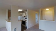 This single-floor residence features an open-concept design that connects the kitchen and living areas, creating a cohesive and functional layout. The kitchen is equipped with essential appliances, including a white refrigerator and an electric range with a stainless steel finish, complemented by a built-in range hood above. Upper cabinetry with warm wood-toned trim provides ample storage, while the under-cabinet lighting adds a practical touch. A raised breakfast bar separates the kitchen from the adjacent living area, offering a casual dining or workspace option. The flooring transitions from dark tile in the kitchen to neutral wall-to-wall carpeting in the living space, clearly defining each zone. A hallway visible in the background leads to additional rooms, suggesting a well-organized floor plan. The textured ceiling and neutral wall tones throughout give the space a clean, understated aesthetic. Ceiling-mounted lighting provides consistent illumination across the main areas. The overall ambiance is practical and comfortable, suited for everyday living.