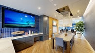 Located at 3300 Boutwell Rd in Lake Worth Beach, Florida, this thoughtfully designed kitchen is a single-level space that maximizes both function and aesthetic appeal. The room is anchored by a striking cobalt blue vertical tile backsplash, which serves as a dramatic backdrop for the built-in wall-mounted flat-screen television and a streamlined wet bar area. Sleek, flat-front gray cabinetry lines the lower wall, paired with crisp marble-look white countertops that provide ample workspace and a clean, modern contrast to the bold tilework. A tall integrated stainless steel refrigerator adds to the polished, contemporary feel. Warm golden-yellow accent walls energize the space and are adorned with large-scale abstract artwork, contributing an artistic, curated character. Light wood-look flooring runs throughout, grounding the design with warmth and continuity. A rectangular dining table with upholstered gray chairs occupies the center of the room, illuminated by a distinctive cascading chandelier with a circular canopy. A textured herringbone-patterned accent wall adds depth and visual interest. Recessed lighting ensures the space is well-lit, while the open sightlines and thoughtful layout make this kitchen both inviting and highly functional.