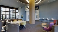 Located at 201 S Elliot Rd in Chapel Hill, NC, this single-floor communal space makes a striking first impression with its soaring ceiling height and dramatic circular overhead lighting fixture that crowns a large cylindrical wood-clad column as its architectural centerpiece. The open-plan layout is thoughtfully organized into distinct seating zones, offering a variety of settings to suit different needs. Collaborative workstations feature clean white surfaces paired with vibrant yellow-green upholstered chairs, while casual lounge areas provide comfortable seating with warm, inviting tones. A bar-height counter along one wall is complemented by sleek white bar stools, ideal for individual work or informal conversation. Contemporary artwork adorns the soft blue-gray walls, adding visual interest and a creative energy throughout the space. Large floor-to-ceiling windows along the exterior wall flood the interior with natural light and offer views of the surrounding streetscape. Patterned carpet grounds the space with texture and warmth, while recessed lighting supplements the natural light for a consistently bright atmosphere. The overall ambiance is modern, dynamic, and welcoming — well-suited for socializing, focused work, or simply relaxing in a thoughtfully designed environment.
