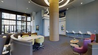 Located at 201 S Elliot Rd in Chapel Hill, NC, this single-floor communal space makes a striking first impression with its soaring ceiling height and dramatic circular overhead lighting fixture that crowns a large cylindrical wood-clad column as its architectural centerpiece. The open-plan layout is thoughtfully organized into distinct seating zones, offering a variety of settings to suit different needs. Collaborative workstations feature clean white surfaces paired with vibrant yellow-green upholstered chairs, while casual lounge areas provide comfortable seating with warm, inviting tones. A bar-height counter along one wall is complemented by sleek white bar stools, ideal for individual work or informal conversation. Contemporary artwork adorns the soft blue-gray walls, adding visual interest and a creative energy throughout the space. Large floor-to-ceiling windows along the exterior wall flood the interior with natural light and offer views of the surrounding streetscape. Patterned carpet grounds the space with texture and warmth, while recessed lighting supplements the natural light for a consistently bright atmosphere. The overall ambiance is modern, dynamic, and welcoming — well-suited for socializing, focused work, or simply relaxing in a thoughtfully designed environment.