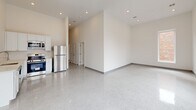Located at 78 Middlesex St in Lowell, MA, this 2-bedroom, 2-bathroom unit offers a clean, modern aesthetic defined by polished terrazzo flooring, crisp white walls, and impressively high ceilings throughout. The open living area is bathed in natural light from a large bank of windows that frame views of the surrounding streetscape and neighboring brick buildings, creating a bright, airy atmosphere. The kitchen is outfitted with stainless steel appliances, including a refrigerator and range, complemented by white cabinetry. A long, well-lit hallway with recessed lighting and a decorative ceiling vent connects the main living spaces to the private quarters. The primary bedroom is generously sized, featuring soaring ceilings, a double closet, and polished terrazzo floors that carry throughout the unit. The second bedroom includes a window with views of the adjacent brick exterior. Both bathrooms are finished in a cohesive white palette, with wall-mounted vanities, LED-backlit mirrors, and clean walk-in shower enclosures. The overall design is minimal and contemporary, with consistent gray baseboard trim and recessed lighting lending a polished, cohesive character to every room.