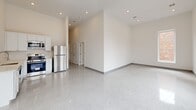 Located at 78 Middlesex St in Lowell, MA, this 2-bedroom, 2-bathroom unit offers a clean, modern aesthetic defined by polished terrazzo flooring, crisp white walls, and impressively high ceilings throughout. The open living area is bathed in natural light from a large bank of windows that frame views of the surrounding streetscape and neighboring brick buildings, creating a bright, airy atmosphere. The kitchen is outfitted with stainless steel appliances, including a refrigerator and range, complemented by white cabinetry. A long, well-lit hallway with recessed lighting and a decorative ceiling vent connects the main living spaces to the private quarters. The primary bedroom is generously sized, featuring soaring ceilings, a double closet, and polished terrazzo floors that carry throughout the unit. The second bedroom includes a window with views of the adjacent brick exterior. Both bathrooms are finished in a cohesive white palette, with wall-mounted vanities, LED-backlit mirrors, and clean walk-in shower enclosures. The overall design is minimal and contemporary, with consistent gray baseboard trim and recessed lighting lending a polished, cohesive character to every room.