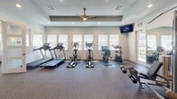 Located at 9102 FM-2920 in Tomball, Texas, this single-floor fitness center is a well-appointed facility designed to support a wide range of workout routines. The space is organized into distinct functional zones, each thoughtfully equipped to meet diverse fitness needs.

The main cardio area features treadmills, elliptical trainers, and stationary bikes, all lined along a wall of windows that flood the room with natural light. A wall-mounted television and ceiling fan with a stylish gold finish enhance comfort during workouts. Recessed lighting and a tray ceiling add a polished architectural touch to this zone.

Adjacent to the cardio area is a strength training section equipped with weight machines, benches, and free weights. Full-length mirrors line the walls, creating a sense of expanded space and allowing users to monitor their form. The room connects seamlessly through interior glass-panel doors, promoting an open and cohesive layout throughout the facility.

A dedicated functional training room offers an organized equipment rack stocked with stability balls, medicine balls, resistance bands, foam rollers, and exercise mats. Multiple windows surround the room, ensuring bright, airy conditions for stretching and group-style training sessions.

Throughout the facility, durable speckled carpet flooring provides a practical and comfortable surface underfoot. The clean, neutral color palette and consistent design language across all areas create a professional and motivating environment for residents and members alike.