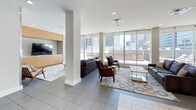 The Sky Lounge is an elevated amenity space designed for both relaxation and social gatherings, offering panoramic views of the surrounding city skyline through expansive floor-to-ceiling glass walls and sliding doors. The interior is thoughtfully organized into several distinct zones. The lounge area features plush leather sofas and accent chairs arranged on a patterned area rug, centered around a glass-top coffee table, with direct access to the outdoor terrace. A built-in media wall with a flat-screen TV adds an entertainment focal point to this seating zone. The games area includes a full-size billiards table beneath a decorative pendant light, providing a fun, social atmosphere. The dining zone accommodates a large rectangular wood table with seating for eight, positioned to take full advantage of the skyline views through the glass walls. The kitchen is well-appointed with a large wraparound bar-height island with ample seating, light wood cabinetry, open shelving against a mosaic tile backsplash, stainless steel appliances including two side-by-side refrigerators, and a microwave. The outdoor terrace extends the entertaining space with lounge chairs and seating arrangements, offering open-air access to sweeping urban views. Light tile flooring throughout ties the space together with a clean, modern aesthetic.