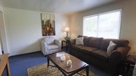 This single-floor residence offers a thoughtfully arranged and inviting living environment. The living room serves as the heart of the space, combining comfort and functionality in equal measure. A large window allows an abundance of natural light to fill the room, creating a bright and airy atmosphere while offering a view of the greenery outside. The seating area is anchored by a generous brown sofa accented with decorative pillows, complemented by a patterned armchair that adds a touch of visual interest. A wooden coffee table sits at the center, providing a practical and stylish focal point for the room. Warm ambient lighting from a classic table lamp lends the space a relaxed and welcoming feel during the evening hours. The carpeted flooring, featuring a woven area rug beneath the seating arrangement, adds texture and warmth underfoot. A piece of colorful abstract wall art hangs prominently, bringing a vibrant artistic element to the otherwise neutral color palette of the walls. The overall ambiance of this space is calm, comfortable, and well-suited for everyday living and relaxed entertaining.