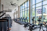 Located at 1901 E Dyer Rd in Santa Ana, California, this contemporary fitness center is designed to deliver a comprehensive workout experience within a thoughtfully appointed, single-level space. The facility is defined by its dramatic, soaring ceilings and expansive floor-to-ceiling glass walls that line the cardio zone, flooding the interior with abundant natural light while offering unobstructed views of a landscaped outdoor courtyard and surrounding community.

The cardio area is equipped with a robust lineup of treadmills, elliptical trainers, stationary bikes, and rowing machines, all positioned to take advantage of the bright, open sightlines. Large industrial-style ceiling fans add a dynamic architectural element while maintaining airflow throughout the space. Adjacent to the cardio section, a dedicated strength training zone features a variety of selectorized weight machines with tan and black upholstery, complementing the gym's modern aesthetic.

A functional training area offers a full rack of medicine balls, TRX suspension trainers, battle ropes, a Bosu ball, and a multi-use rig for versatile, high-intensity workouts. A separate strength room houses power racks with barbells and weight plates, accented by bold motivational wall typography. The entry is framed by sleek glass doors with a branded frosted graphic, providing a polished welcome into this well-equipped fitness facility.