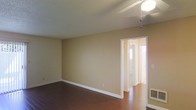 This single-floor space presents a straightforward and functional layout finished with warm, neutral-toned walls that lend a welcoming ambiance throughout. The main living area is open and uncluttered, featuring rich hardwood flooring that adds warmth and visual depth underfoot. White baseboards provide a crisp contrast against the beige walls, contributing to a clean and cohesive aesthetic. A ceiling fan with a light fixture is centrally positioned overhead, offering both ventilation and ambient lighting. The textured ceiling adds a subtle architectural detail to the space. A doorway with white trim leads into an adjoining room or hallway, suggesting a connected layout that allows for easy flow between areas. Natural light filters in from the adjacent space, brightening the interior. A wall-mounted vent near the floor indicates a central heating and cooling system. Overall, the space is neutral and move-in ready, offering a blank canvas that adapts easily to a variety of furnishing styles and interior preferences.