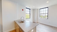 Located at 1 Atlantic St in downtown Stamford, Connecticut, this single-floor unit showcases a clean, contemporary design throughout. The interior is unified by light hardwood-style flooring and crisp white walls, creating a bright and cohesive aesthetic from room to room.

The kitchen serves as a central feature of the living area, anchored by a peninsula with a light wood base and smooth quartz countertop that houses a stainless steel undermount sink. The kitchen is fully equipped with stainless steel appliances, including a refrigerator, range, microwave, and dishwasher, complemented by warm wood cabinetry with under-cabinet lighting. Large, black-framed grid-pane windows flood the open living and kitchen area with natural light while offering views of the surrounding cityscape and tree canopy.

A hallway connects the living spaces to the private rooms, lined with panel-style white doors accented by matte black hardware. The bedroom features its own generously sized grid-pane window, continuing the natural light theme. The bathroom is finished with white subway tile surrounding a soaking tub and shower combination, with gray tile flooring providing a subtle contrast. Recessed lighting throughout the unit ensures consistent, even illumination across all spaces.