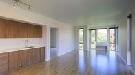 This single-floor unit showcases a thoughtfully designed open-concept layout that seamlessly connects the kitchen and living areas. The warm-toned hardwood flooring runs continuously throughout, lending a sense of cohesion and spaciousness to the interior. The kitchen is outfitted with rich wood cabinetry offering ample storage above and below a clean countertop, complemented by a simple, functional sink. The cabinetry's warm brown tones contrast nicely against the crisp white walls, creating a balanced aesthetic.

The living area extends generously toward a striking wall of floor-to-ceiling windows that flood the space with abundant natural light. These expansive windows frame lush, verdant tree views, bringing the outdoors in and enhancing the unit's airy ambiance. A wall-mounted climate control unit is positioned beneath the windows for added comfort.

A doorway off the main living area leads to an additional room, suggesting flexible use for sleeping or storage. The smooth white ceilings and clean wall finishes throughout contribute to a modern, understated aesthetic that is both practical and inviting.