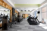 This contemporary fitness center is thoughtfully designed to support a full range of workout routines in a bright, open environment. The space is anchored by floor-to-ceiling windows along its exterior walls, flooding the interior with natural light and offering views of the surrounding urban streetscape. Light-toned, wood-grain luxury vinyl plank flooring runs throughout, complementing the clean white ceilings accented with lime-green trim details and industrial-style cage pendant fans.

The cardio zone is well-appointed with multiple treadmills, a StairMaster, an elliptical trainer, and upright stationary bikes, all arranged to take advantage of the natural light and outdoor views. A dedicated cycling area features indoor spin bikes positioned in front of a large wall mirror framed by a striking textured wood-panel accent wall, creating an immersive workout zone. The strength training section offers cable machines, a multi-functional weight rack, dumbbells, and a weight bench, all set against the same decorative wood-tile accent wall with large mirrors that amplify the sense of space.

A separate stretching and functional training area is visible through glass-panel sliding doors and features stability balls and open floor space. Wall-mounted televisions are positioned throughout for entertainment. The overall ambiance is modern, energetic, and welcoming, combining high-end finishes with practical, professional-grade fitness equipment.
