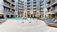 Located in Charlotte's vibrant NoDa neighborhood, this impressive amenity level at 2226 North Davidson Street offers a seamless blend of indoor comfort and outdoor living across a generously sized single-floor footprint. The space is anchored by a striking circular outdoor pool surrounded by lounge chairs and umbrella-shaded seating, set within a beautifully landscaped courtyard framed by the building's multi-story brick and contemporary facade. String lights and planter boxes with seasonal flowers add warmth to the al fresco dining and grilling area nearby. Inside, the space transitions fluidly from the pool deck through large-pane glass doors into a covered indoor lounge area featuring coffered ceilings, warm wood built-in benches with decorative tile detailing, and an eye-catching living green wall. Egg-shaped hanging chairs provide cozy nooks beside the verdant backdrop. Additional interior zones include a turf-floored social lounge with a whitewashed brick accent wall and mounted TV, package lockers, and planter-lined seating areas bathed in natural light from oversized windows. Throughout, the design combines natural materials — wood, stone, and greenery — with clean white architectural lines to create an inviting, resort-inspired atmosphere ideal for relaxation and community gathering.