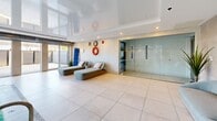 Located at 17 Place de la Triade in Pointe-Claire, Quebec, this single-floor indoor pool facility offers a sophisticated and tranquil aquatic environment. The centerpiece of the space is a generously sized indoor swimming pool with crystal-clear turquoise water, illuminated from below to create a vivid, inviting atmosphere. Stainless steel pool entry rails provide safe and elegant access to the water.

The pool is framed by wide-format tile flooring in neutral tones, contributing to the clean, contemporary aesthetic throughout. Along one side of the pool, a raised lounging terrace features stylish seating — including sleek chairs and a soft curved sofa — accompanied by lush tropical plants that add warmth and a resort-like ambiance.

A glossy, high-sheen ceiling reflects light beautifully across the space, amplifying the sense of openness and airiness. Floor-to-ceiling sliding glass doors and expansive windows line one wall, flooding the interior with natural light while offering views of the outdoor surroundings. A dedicated relaxation area near the entrance includes modern chaise lounges, with glass-partitioned rooms nearby providing additional amenity access. Safety equipment is discreetly mounted on the walls. Overall, this space combines functional design with upscale finishing for a premium recreational experience.