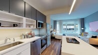 Located at 165 Huguenot St in New Rochelle, NY, this contemporary studio offers a thoughtfully designed open-plan layout that seamlessly integrates the living, sleeping, and dining areas into a single, cohesive space. Warm hardwood flooring flows throughout, complementing the modern aesthetic and creating a welcoming ambiance.

The well-appointed kitchen runs along one wall and is outfitted with stainless steel appliances, including a range, dishwasher, microwave, and full-size refrigerator. Sleek dark upper cabinetry contrasts with crisp white lower cabinets and open shelving, while a clean white countertop and subway tile backsplash add polish. A white kitchen island with bar seating provides additional prep space and a casual dining area.

The living and sleeping area is bright and airy, benefiting from a large window that invites generous natural light and offers outdoor views of the surrounding neighborhood. Recessed linear lighting above adds a modern touch to the ceiling design.

The bathroom is clean and efficiently designed, featuring a white vanity with under-cabinet storage, a framed mirror, and light tile flooring, maintaining the unit's consistent modern and minimalist style throughout. This studio is a refined urban residence ideal for comfortable single-occupancy living in the heart of New Rochelle.