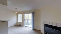 This single-floor residence offers a welcoming and functional layout filled with natural light. The living area serves as the heart of the space, featuring soft carpeted flooring and warm neutral-toned walls that contribute to a calm, understated ambiance. A sliding glass door opens directly onto a private balcony, framing pleasant views of surrounding greenery and mature trees, while an adjacent window with horizontal blinds further enhances the sense of openness and connection to the outdoors. A baseboard heater beneath the window provides efficient climate control. A tile-surround fireplace anchors one end of the living area, offering both a functional heating element and a visual focal point. Overhead, a decorative pendant light fixture adds a touch of character to the space. The textured ceiling completes the room's classic residential aesthetic. Overall, the space presents a straightforward, comfortable living environment with practical features, natural light, and easy outdoor access — ideal for those seeking a peaceful, low-maintenance home.