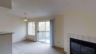 This single-floor residence offers a welcoming and functional layout filled with natural light. The living area serves as the heart of the space, featuring soft carpeted flooring and warm neutral-toned walls that contribute to a calm, understated ambiance. A sliding glass door opens directly onto a private balcony, framing pleasant views of surrounding greenery and mature trees, while an adjacent window with horizontal blinds further enhances the sense of openness and connection to the outdoors. A baseboard heater beneath the window provides efficient climate control. A tile-surround fireplace anchors one end of the living area, offering both a functional heating element and a visual focal point. Overhead, a decorative pendant light fixture adds a touch of character to the space. The textured ceiling completes the room's classic residential aesthetic. Overall, the space presents a straightforward, comfortable living environment with practical features, natural light, and easy outdoor access — ideal for those seeking a peaceful, low-maintenance home.