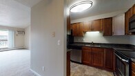 This single-floor space is thoughtfully designed to maximize functionality and comfort across its key areas. The kitchen serves as the centerpiece of the unit, showcasing a U-shaped layout with rich, medium-toned wood cabinetry extending from floor to ceiling, providing generous storage throughout. Dark granite countertops with subtle veining offer a striking contrast against the cabinetry and are paired with a full suite of stainless steel appliances, including a refrigerator, dishwasher, electric range, and over-the-range microwave. Under-cabinet lighting illuminates the workspace, while stone-look tile flooring adds durability and visual warmth. A flush-mount ceiling fixture provides ample overhead lighting. The kitchen is accessible from an adjacent hallway, which transitions seamlessly between rooms. The bathroom features a full tub-shower combination with cream-toned tile surround, a vanity with dark wood cabinetry, a white countertop, and matching stone-look tile flooring, creating a cohesive aesthetic. The bedroom is a calm, neutral space with soft carpeting, light gray walls, white panel doors, and clean white baseboards, offering a comfortable and versatile environment.