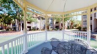 Gazebo 2 is a single-level community amenity space combining two distinct areas: an open-air gazebo and a modern fitness center. The gazebo features classic architectural detailing, including white-painted spindle railings, decorative columns, and a gently curved perimeter that offers panoramic views of the surrounding landscaped courtyard. Wrought iron seating provides a comfortable spot to relax outdoors, while mature trees and well-maintained grounds create a tranquil backdrop.

The fitness center is bright and thoughtfully designed, with neutral-toned walls, recessed lighting, and ceiling fans that promote air circulation throughout the space. Floor-to-ceiling mirrors line the walls, visually expanding the room and allowing users to monitor their form during workouts. Cardio equipment includes multiple treadmills and stationary bikes, while a comprehensive free weight section offers a full dumbbell rack and medicine balls in various weights. Strength training is further supported by cable machines and an adjustable bench press station. Wooden open shelving provides organized storage for rolled towels, fitness accessories, and decorative greenery. Large sliding glass doors and windows flood the space with natural light and offer views of the outdoor pool area, creating a seamless connection between the interior and exterior environments.