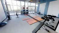 This single-floor fitness center offers a thoughtfully designed workout environment filled with an array of exercise equipment to support a variety of training styles. The space is anchored by large, floor-to-ceiling windows framed in dark steel, allowing abundant natural light to pour in and providing open views of the surrounding exterior — creating an energizing atmosphere for exercise. The light grey flooring provides a clean, neutral base throughout the room. A full-length mirror lines one wall, enhancing the sense of space and allowing users to monitor their form during workouts. Equipment includes a rowing machine, an air bike, and a weight bench, catering to both cardio and strength training needs. Exercise mats are laid out across the central floor area, offering dedicated space for stretching, yoga, or floor-based workouts. A rack of stability balls in varying sizes adds versatility for functional fitness routines. Pendant lighting in muted tones complements the cool blue accent wall, contributing to a focused yet modern aesthetic. The overall ambiance is clean, open, and purposefully organized, making the space well-suited for a range of fitness activities.
