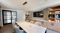 This sophisticated conference room offers a well-appointed space designed for productive meetings and collaborative work. At its center sits a substantial stone-topped conference table with integrated power access, surrounded by comfortable upholstered chairs that accommodate a full team. A large wall-mounted flat-screen display serves as the focal point on one wall, flanked by warm-toned built-in cabinetry and open shelving that spans the full width of the room. The shelves are thoughtfully styled with books and decorative accents, lending the space a refined, professional character. Geometric pendant lighting in a modern linear design hangs above the table, providing focused illumination that complements recessed ceiling lights throughout. The room is enclosed by floor-to-ceiling glass partition walls with bold black steel frames, offering a sense of openness and visual connectivity to the surrounding space while maintaining privacy. Natural light filters in through exterior windows, brightening the interior and enhancing its airy feel. Underfoot, a patterned carpet in a subtle houndstooth design adds texture and warmth to the overall aesthetic. The combination of contemporary finishes, functional built-ins, and transparent boundaries makes this conference room a standout space for professional collaboration.