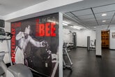 Located at Huntington Village in Huntington Beach, California, this well-appointed fitness center spans a generously sized single-floor layout divided into three distinct functional areas. The main workout area makes an immediate impression with its bold, large-scale motivational mural — a striking boxing-themed graphic with the phrase "Sting Like a Bee" — setting an energetic tone throughout the space. This primary area houses a variety of cardio equipment, including ellipticals and upright bikes, and is complemented by a wall-mounted flat-screen TV. Overhead, bright linear fluorescent lighting ensures the space is thoroughly illuminated. An adjacent strength training room provides dedicated space for resistance machines and cable equipment, with clean white walls, dark rubber flooring, and a glass-paneled door for separation while maintaining an open feel. A third room offers additional cardio equipment, including treadmills and stair climbers, and benefits from multiple windows that bring in natural light and provide views of the surrounding outdoor pool and courtyard area. The entry area features dual water fountains, a bench seating area, and glass doors leading to the exterior stairway and outdoor grounds. Throughout, the design maintains a consistent modern, clean aesthetic with a neutral color palette, dark durable flooring, and efficient overhead lighting.