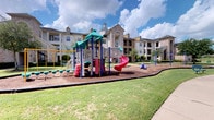 This residential community amenity space showcases a thoughtfully designed leasing office and clubhouse building, characterized by a striking exterior that combines white lap siding with natural stone accent pillars. The entrance features a dramatic arched window with floor-to-ceiling glass panes, allowing abundant natural light to pour into the interior while offering views of the surrounding community. A covered walkway with substantial stone columns leads visitors along a smooth concrete path to the main entry, creating an inviting and sheltered arrival experience.

The outdoor amenities are a standout highlight of this community. A generously sized resort-style swimming pool takes center stage, featuring a freeform design and a decorative stone water feature surrounded by tall, swaying palm trees. The expansive pool deck is finished in textured concrete, providing ample space for lounging and outdoor enjoyment under open skies.

Adjacent to the residential buildings, a vibrant children's playground offers a colorful multi-level play structure complete with slides, climbing elements, and stepping features, all set on a cushioned wood chip surface bordered by manicured green lawns. The multi-story residential buildings surrounding these amenities feature balconies, arched architectural details, and a warm mix of stone and stucco finishes, contributing to the cohesive and upscale aesthetic of the overall community.