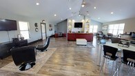 This single-floor space offers an open, airy layout anchored by striking vaulted ceilings that lend a sense of grandeur and spaciousness throughout. The warm wood-look flooring runs seamlessly across the entire area, complementing the clean, modern aesthetic. Recessed lighting and ceiling-mounted pendant fixtures illuminate the space evenly, while the high ceiling line enhances the overall sense of openness.

The main lounge area features a bold red sectional sofa paired with a white tufted ottoman, centered on a patterned area rug. A wall-mounted flat-screen television is flanked by built-in shelving niches with integrated lighting, creating a stylish and functional focal point. Decorative items displayed in the niches add character and warmth to the environment.

Toward the front of the space, a counter-height bar table with modern swivel chairs provides an alternative seating option, ideal for casual conversation or individual use. Toward the rear, additional seating areas offer flexible arrangements for a variety of gathering needs. Vertical shiplap-style wall paneling adds a contemporary architectural detail that runs along key interior walls, tying the design together. Large windows allow natural light to filter into the space, contributing to an inviting and well-lit atmosphere throughout.