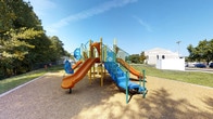 This outdoor playground features a colorful, multi-level play structure designed to encourage active play and physical development. The structure is built with a combination of bold orange, blue, and yellow components, creating an eye-catching and inviting environment for children. Two curved orange slides extend from elevated platforms, offering exciting descent options, while blue staircases and railings provide safe access to the upper levels. The play structure also incorporates climbing panels and various interactive elements along its framework, adding variety to the play experience. The surrounding ground is covered with a wood chip safety surface, providing cushioning beneath the equipment. The playground is set within a spacious open area with neatly maintained grass surroundings and mature trees lining the perimeter, offering natural shade. A small utility structure is visible nearby, and a fence encloses part of the background area. The setting benefits from an abundance of natural light under a clear blue sky, making it a bright and welcoming recreational space suited for children of various ages.