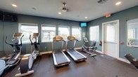 This single-level fitness center is designed to support a comprehensive workout experience, combining cardio and strength training areas within one cohesive space. The interior is characterized by teal-painted walls, recessed lighting, and durable rubber flooring that provides both safety and comfort underfoot.

The cardio zone is positioned along a windowed wall, offering views of the outdoor surroundings. It is equipped with multiple treadmills, elliptical trainers, and a recumbent bike, each oriented to take advantage of the natural light streaming through the windows. Wall-mounted flat-screen televisions are installed throughout, allowing users to stay entertained during workouts.

The strength training area features a selection of cable and selectorized weight machines, a dedicated free weight section with a full dumbbell rack, and an adjustable workout bench. Large wall mirrors span much of the interior, visually expanding the space and allowing users to monitor their form.

Additional amenities include a water cooler station and convenient access to adjacent areas through multiple doorways. The overall ambiance is clean, functional, and well-organized, making this fitness center a practical and inviting space for a wide range of exercise routines.
