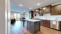 The Tiller floor plan at Carrick Tonka Bay is a thoughtfully designed two-story residence offering two bedrooms and two and a half bathrooms. The main living level features rich hardwood-style flooring throughout, anchoring an open-concept layout that connects the kitchen, dining, and living areas. The kitchen is a standout, showcasing warm walnut-toned cabinetry, a white subway tile backsplash, stainless steel appliances including double wall ovens, a gas cooktop, and a central island with contrasting cabinetry and white quartz countertops. The living room is bright and inviting, with large windows and glass doors opening to a balcony, alongside a fireplace that adds a focal point to the space. A dedicated dining area with hardwood-style flooring offers a comfortable setting for everyday meals. Upstairs, both bedrooms are carpeted and filled with natural light from generously sized windows. The primary suite includes a spa-inspired en-suite bathroom featuring a dual-sink vanity with dark wood cabinetry, quartz countertops, and a large framed mirror. Walk-in closet space with wire shelving provides ample storage. A stacked in-unit washer and dryer adds everyday convenience to this well-rounded residence.
