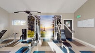This modern fitness center is thoughtfully designed to support a comprehensive workout experience within a single, open-plan space. The room is distinguished by soaring vaulted ceilings accented by recessed lighting and ceiling fans, creating an airy, energizing atmosphere throughout.

The flooring features a bold geometric pattern in shades of teal, gray, and neutral tones, lending a contemporary visual appeal underfoot. The cardio zone is well-appointed with multiple treadmills and an elliptical machine, positioned near frosted windows that allow natural light to filter in while maintaining privacy. A glass door provides direct access to an outdoor area, visible as a pool beyond.

The strength training section offers a multi-station cable machine, weight bench, and a functional training rack complete with TRX suspension straps, kettlebells, medicine balls, and resistance equipment. A large wall-mounted mirror visually expands the space and supports form-focused training. A vibrant floor-to-ceiling photographic mural depicting a serene outdoor yoga scene serves as a striking focal point, reinforcing a wellness-oriented ambiance.

Additional amenities include a wall-mounted flat-screen television, a water dispenser, and an open arched passageway connecting sections of the facility. The neutral taupe walls and clean white trim contribute to the space's polished, welcoming aesthetic.