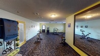 Located at 7135 Islander Dr in Indianapolis, Indiana, this single-floor fitness center offers a comprehensive workout environment designed to accommodate a variety of exercise routines. The space is finished with durable, patterned carpet flooring and textured ceilings, giving it a practical and functional feel throughout.

The main workout area is spacious and open, featuring light green and yellow accent walls that create an energizing atmosphere. Cardio equipment includes a treadmill, elliptical trainer, and stationary cycling bike, all arranged to allow comfortable spacing between users. A weight bench and a dedicated free weights section house a full rack of dumbbells, catering to strength training needs. A large wall-mounted mirror in the free weights area enhances the sense of space and allows for form checking during workouts.

A separate alcove area contains a cable resistance machine, providing additional strength and functional training options. Motivational artwork and posted fitness center rules are displayed on the walls, contributing to a focused and professional environment. The space is accessed through a yellow-trimmed entry door and is equipped with adequate overhead lighting and ventilation. Exercise balls and other fitness accessories round out the available equipment, making this fitness center a well-equipped facility for residents and users of all fitness levels.