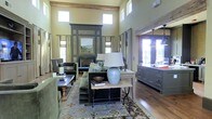 This single-story residence showcases an impressive open-concept design defined by soaring ceilings and exposed wooden ceiling beams that lend a rustic yet refined character to the interior. The living area is generously proportioned, anchored by a bold, dark-framed fireplace surround that serves as a dramatic focal point. Rows of clerestory windows positioned high along the walls flood the space with natural light while maintaining privacy, complementing the warm, sage-green walls and rich hardwood flooring throughout.

The living room is thoughtfully furnished with plush, earth-toned seating arranged around a patterned area rug, creating an inviting and cohesive gathering space. A reclaimed wood console table and wrought-iron accents add to the space's artisan aesthetic. Tall linen drapes frame the windows, softening the architectural lines and adding warmth to the room.

Adjacent to the living area, the kitchen is visible through an open passageway framed by a rustic wood beam. It features dark sage cabinetry with vintage-style pendant lighting, offering a seamless continuation of the home's natural color palette. The open flow between spaces makes the layout ideal for entertaining, while the consistent use of natural materials and muted tones creates a cohesive, welcoming ambiance throughout.