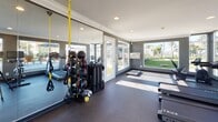 This single-floor fitness center is thoughtfully designed to support a full range of workout activities in a bright, modern environment. The space is finished with dark rubber flooring, crown molding, and recessed lighting, giving it a polished, professional aesthetic. Large picture windows and glass doors wrap around the room, flooding the interior with natural light and offering expansive views of a lush artificial turf lawn and an outdoor pool area with seating — creating an energizing atmosphere for exercise.

The facility is comprehensively equipped for both cardio and strength training. Cardio machines include treadmills and a stationary upright bike, while strength training options encompass a multi-station cable machine, a full dumbbell rack with a variety of weights, an adjustable workout bench, and a dedicated TRX suspension training system with resistance bands and medicine balls on a central rack. Full-length mirrors line one wall, visually expanding the space and allowing users to monitor their form.

A wall-mounted flat-screen television adds a multimedia element to the workout experience. An exterior door provides direct access to the outdoor amenity area, seamlessly connecting the indoor fitness space with the surrounding recreational grounds. Overall, the fitness center offers a well-rounded, light-filled environment suited for diverse training needs.