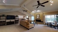 This single-story space makes a strong impression with its dramatic vaulted ceilings accented by exposed white beams that draw the eye upward, creating a sense of generous vertical space throughout the main living area. Two ceiling fans are strategically positioned to circulate air across the open floor plan, complementing the track lighting that provides adjustable illumination. The living room serves as the centerpiece, anchored by a full brick fireplace with a classic hearth, adding warmth and a traditional architectural focal point. A framed artwork piece above the mantel enhances the refined yet comfortable aesthetic. The living area features plush carpeting underfoot and is furnished with a generous sectional sofa and glass-topped coffee table, emphasizing the spacious, open feel. Large windows allow natural light to filter into the space. The kitchen is visible from the living room, separated by a raised bar counter, and is equipped with white cabinetry and appliances, maintaining a clean and functional design. Crown molding throughout adds a polished finishing detail. The overall ambiance is warm and inviting, blending casual comfort with well-considered architectural elements.