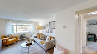 This single-floor residence offers a thoughtfully arranged layout that balances comfort and functionality across its key living areas. The living room serves as the heart of the home, featuring soft carpet flooring, a textured ceiling, and a large window that fills the space with natural light and offers views of the surrounding greenery. The room accommodates a generous seating arrangement with ample space for both relaxing and entertaining. The kitchen is efficiently designed with light wood cabinetry, granite-style countertops, and black appliances including a gas range, over-the-range microwave, and a full-size refrigerator. Durable wood-look flooring adds a modern touch to this functional space. The primary bedroom is a serene retreat, finished with plush carpet, sheer window dressings, and a wall-mounted air conditioning unit, ensuring year-round comfort. A second bedroom offers similar finishes with bright natural light and a flexible layout suitable for a guest room or office. The bathroom is clean and classic, featuring white tilework, a mirrored medicine cabinet, and a shower-tub combination. Throughout the residence, a neutral color palette and consistent design elements create a cohesive and inviting atmosphere.