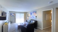 This single-floor living space centers around a comfortable, well-appointed living room designed for both relaxation and everyday use. The room features plush carpeted flooring that extends throughout, adding warmth underfoot. A large sliding glass door fitted with vertical blinds and sheer curtains serves as the focal point of one wall, drawing in generous amounts of natural light and offering a view of the greenery outside.

The seating area is anchored by a dark leather sofa accented with light blue throw pillows, paired with a matching armchair and a warm-toned wooden coffee table. Two tall floor lamps flank the sofa, providing ambient lighting and a sense of symmetry. The walls are painted in a soft gray tone, offering a neutral backdrop that complements the vibrant, colorful abstract artwork displayed prominently above the sofa and on an adjacent wall, adding personality and visual interest to the space.

A doorway visible to the right suggests easy access to additional areas of the unit. The textured white ceiling and clean baseboards contribute to a tidy, well-maintained aesthetic throughout. Overall, the space feels bright, inviting, and thoughtfully arranged.