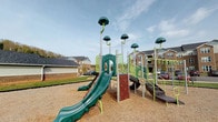 This single-story community amenity facility offers a thoughtfully designed set of spaces catering to residents of all ages. The interior is finished with neutral, light-toned walls, white trim, and durable speckled flooring throughout, creating a clean and cohesive atmosphere. A carpeted corridor with glass-paneled doors connects the various areas, lending an open and accessible feel to the layout.

The fitness center is generously proportioned and well-lit by multiple windows dressed with white blinds, allowing ample natural light to fill the room. It is equipped with cardio machines including ellipticals, stationary bikes, and a variety of free weights, stability balls, and strength equipment, providing a comprehensive workout environment.

Adjacent to the fitness area is a dedicated children's play nook, separated by a gated half-wall for safety. The space features colorful toys, a play tent, and activity rugs, offering a secure and engaging area for young residents.

A bright, tile-floored corridor connects to additional community rooms, adding a sense of depth and flow to the facility. Outside, a multi-level playground structure with slides, climbing panels, and whimsical mushroom-topped poles sits on a rubberized surface, surrounded by views of the broader residential community.