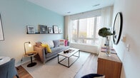 Located at 165 Huguenot Street in New Rochelle, NY, this single-floor residence offers a well-designed, contemporary living experience. The open-concept layout connects the living, dining, and kitchen areas seamlessly, creating a cohesive and functional flow throughout the space.

The living area is distinguished by soft seafoam-blue accent walls, warm hardwood-style flooring, and a large picture window that invites abundant natural light and urban views. A picture ledge display shelf adds a curated, gallery-like touch above a comfortable seating arrangement centered around a sleek coffee table.

The kitchen is thoughtfully appointed with two-tone cabinetry — crisp white lower cabinets paired with charcoal upper cabinets — stainless steel appliances, open display shelving, and a white waterfall-edge island with bar seating, ideal for casual dining or meal prep. Adjacent to the kitchen, a compact dining nook provides additional seating for everyday meals.

The bedroom is a serene retreat, featuring a large window with city views, soft neutral tones, built-in closet storage, and warm accent lighting. The bathroom presents a clean, minimalist aesthetic with light tile flooring, a white vanity with an undermount sink, and a large frameless mirror. Throughout the residence, modern finishes and a neutral palette create a polished and inviting atmosphere.