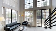 This sophisticated two-story space makes an immediate impression with its dramatic double-height living area, where floor-to-ceiling grid-pane windows stretch upward to flood the interior with natural light. The towering glazed walls offer sweeping views of lush palm trees and the surrounding outdoor environment, creating a seamless connection between inside and out. The main living area is anchored by sleek black leather seating arranged around a minimalist coffee table, complementing the clean, neutral palette of white walls and light carpeting. A bold, colorful sculptural floor lamp adds an artistic accent, reinforcing the modern aesthetic. Large sliding glass doors open directly to an outdoor area, enhancing the sense of openness and encouraging indoor-outdoor living. The architectural design is distinctly contemporary, characterized by clean lines, a restrained color scheme, and an emphasis on volume and light. The upper level overlooks the main living space, contributing to the airy, open feel of the interior. Throughout, the design prioritizes spaciousness, natural illumination, and a refined modern sensibility that creates an inviting yet architecturally compelling environment.