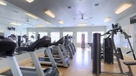 This fitness center is a thoughtfully designed, single-floor facility built to support a comprehensive workout experience. The space is outfitted with a row of cardio machines, including treadmills and ellipticals, arranged to maximize both usability and flow. Strength training equipment and a punching bag round out the workout offerings, catering to a variety of fitness routines. Large windows line the walls, flooding the room with natural light and creating an open, energizing atmosphere. Full-length mirrors span an entire wall, enhancing the sense of space while allowing users to monitor their form during exercise. The light wood-toned flooring adds warmth to the interior, complementing the clean, neutral-toned walls. A ceiling fan overhead provides additional airflow and comfort during workouts. Recessed and flush-mounted lighting fixtures ensure the space remains bright and well-lit at all times. The overall layout is clean, organized, and efficient, making this fitness center a practical and welcoming environment for users of all fitness levels.
