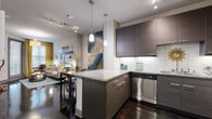 This single-floor residence showcases a cohesive modern design throughout its open layout. The kitchen is a standout feature, equipped with stainless steel appliances including a dishwasher, electric range, and refrigerator. Dark, flat-front cabinetry extends to the ceiling, complemented by a glass tile backsplash, granite countertops, and pendant lighting that hangs above the central island. Polished concrete floors run throughout the space, lending an urban, industrial aesthetic that ties each area together.

Adjacent to the kitchen, the dining area accommodates an intimate table-and-chair set beneath coordinating pendant lights, with bold artwork adding a vibrant focal point. The living room is bright and welcoming, featuring large windows dressed with patterned curtains, track lighting, and ample wall space for oversized art.

The bedroom is a comfortable retreat with room for a large bed, multiple windows allowing natural light, and coordinating decor. The bathroom features a full soaking tub with a tiled surround, granite-topped vanity with dark cabinetry, polished concrete flooring, and a curved shower rod—maintaining the modern aesthetic carried throughout the entire space.
