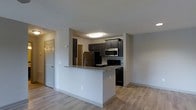 This single-floor residence offers a thoughtfully designed open-concept layout that seamlessly connects the kitchen and living areas, creating an inviting and functional living environment. The kitchen is a standout feature, showcasing dark espresso-toned cabinetry that provides generous storage, complemented by a granite-topped peninsula that doubles as a breakfast bar and prep space. Black and stainless steel appliances — including a refrigerator, range, and microwave — lend a sleek, contemporary aesthetic. Light hardwood-style flooring runs throughout the space, adding warmth and visual continuity between rooms. The neutral, light grey wall tones create a clean and versatile backdrop that adapts well to a variety of décor styles. Flush-mount ceiling lighting provides consistent illumination across the open area. A textured ceiling adds subtle architectural character throughout. Doorways visible from the main living space suggest easy access to additional rooms, contributing to a practical and well-connected floor plan. Overall, the space strikes a balance between modern design sensibilities and comfortable everyday living, making it an accessible and attractive single-level home.
