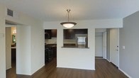 This single-floor residence offers a thoughtfully designed open-concept layout that seamlessly connects the living and kitchen areas. Rich, dark hardwood flooring runs throughout, adding warmth and a sense of continuity to the space. The kitchen is well-appointed with dark espresso cabinetry, a stainless steel refrigerator, a black gas range, and a granite-topped pass-through counter that opens toward the living area — ideal for both cooking and entertaining. A stylish pendant light with a chain-hung bowl fixture hangs above the pass-through, serving as a central focal point that ties the spaces together. The kitchen features ample upper cabinet storage and a sink positioned beneath additional cabinetry. The living area is bright and open, with neutral white walls that complement the darker tones of the flooring and cabinetry. A doorway visible from the main living space leads to what appears to be a walk-in closet or additional storage area, hinting at practical storage solutions throughout. The textured ceiling adds a subtle design detail overhead. Overall, the space presents a clean, modern aesthetic with a functional and comfortable layout suited for everyday living.